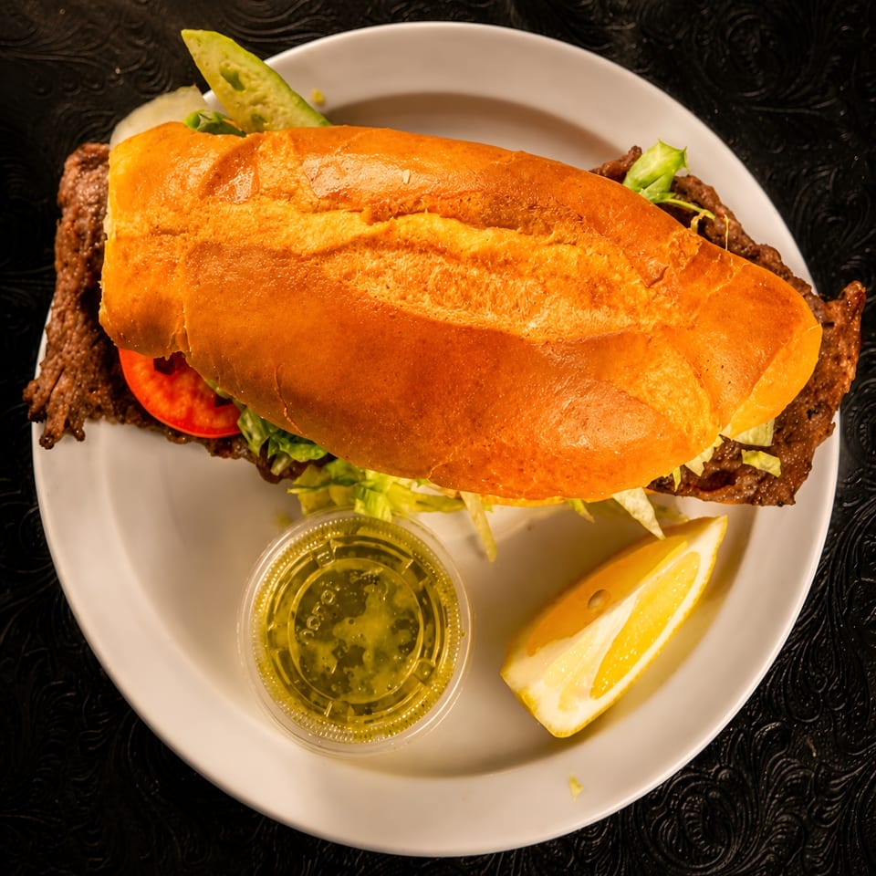 A large sandwich with meat, lettuce, and tomato on a white plate with a lemon wedge and sauce.