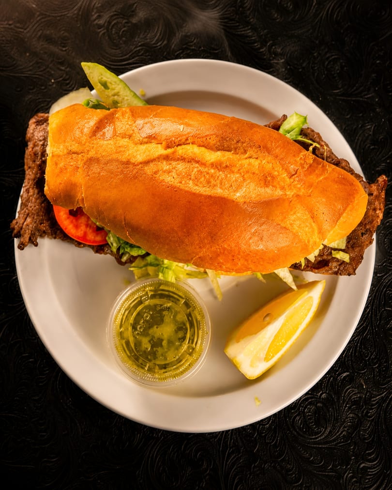 A large sandwich with meat, lettuce, and tomato on a white plate with a lemon wedge and sauce.