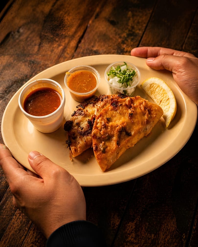 Hands holding a plate with two golden-brown quesadillas, two sauces, chopped onions, and a lemon wedge.